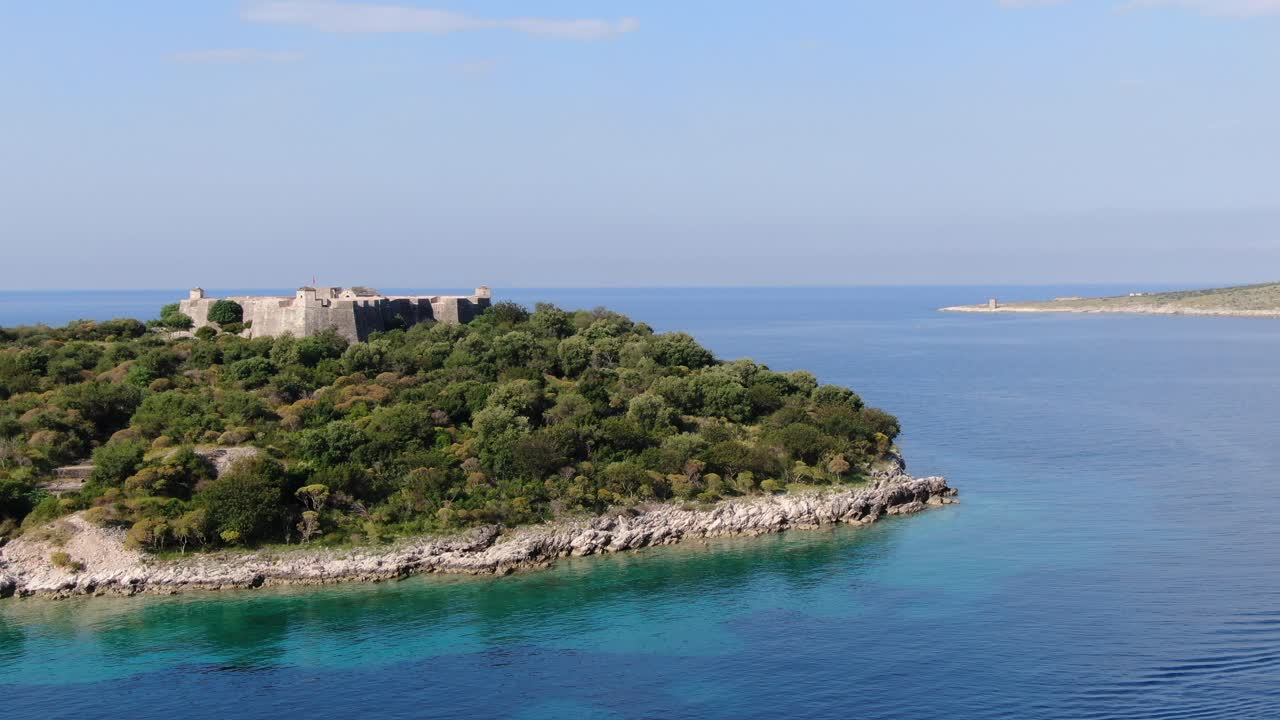 알바니아에서 드론이 푸른 은 바다 위를 날아다니며 은 날 중세 요새가 있는 작은 초록색 섬을 볼 수 있습니다.