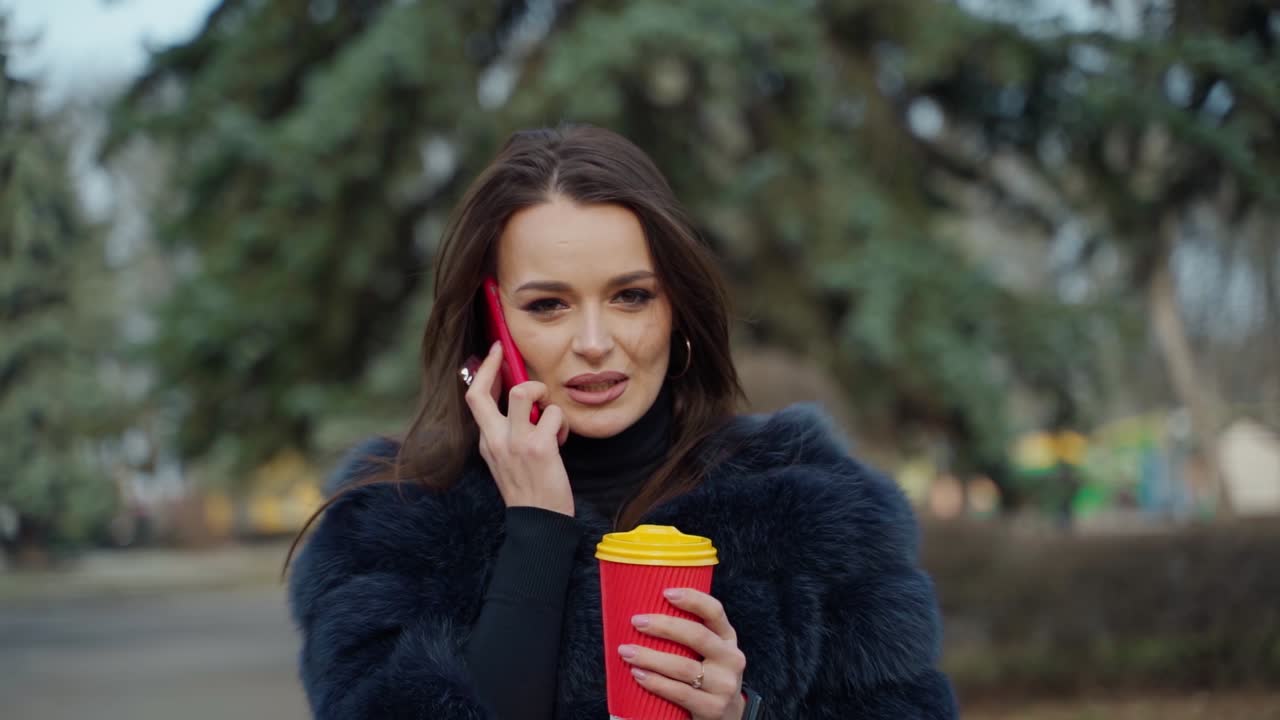 Girl in fur coat with a phone and coffee outdoors. Portrait of a young woman talking the mobile phone and drinking hot beverage in the park. Slow motion.