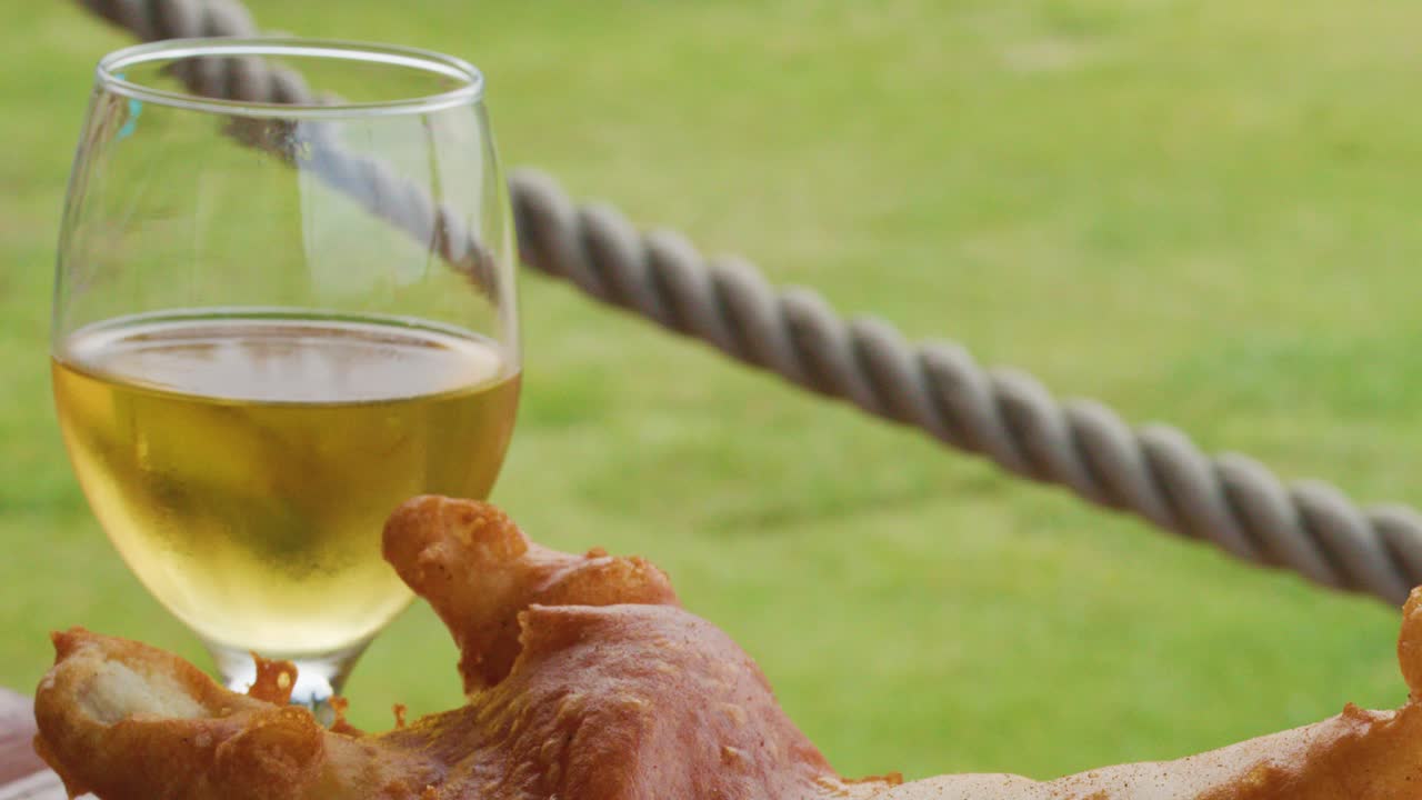 A glass of blonde beer is poured beside crispy fish and chips on an outdoor table, with natural daylight and a grassy background