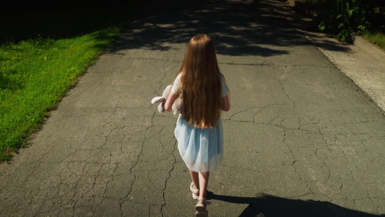 Back view of young girl walking alone on cracked sunlit road while holding stuffed animal, moving toward shaded part of pavement lined with grass and curbs under bright daylight
