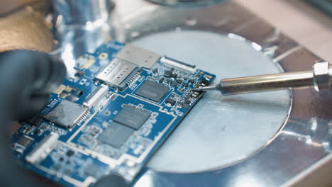 a close-up view of a technician's gloved hands using a soldering iron to work on a blue circuit board under a microscope