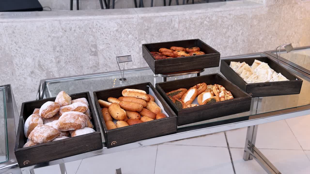 Tavolo buffet per la colazione con panini freschi assortiti, ciambelle zuccherate, pasticcini salati ripieni e focaccia a fette, disposti in vassoi di legno rustici su un espositore di vetro all'interno