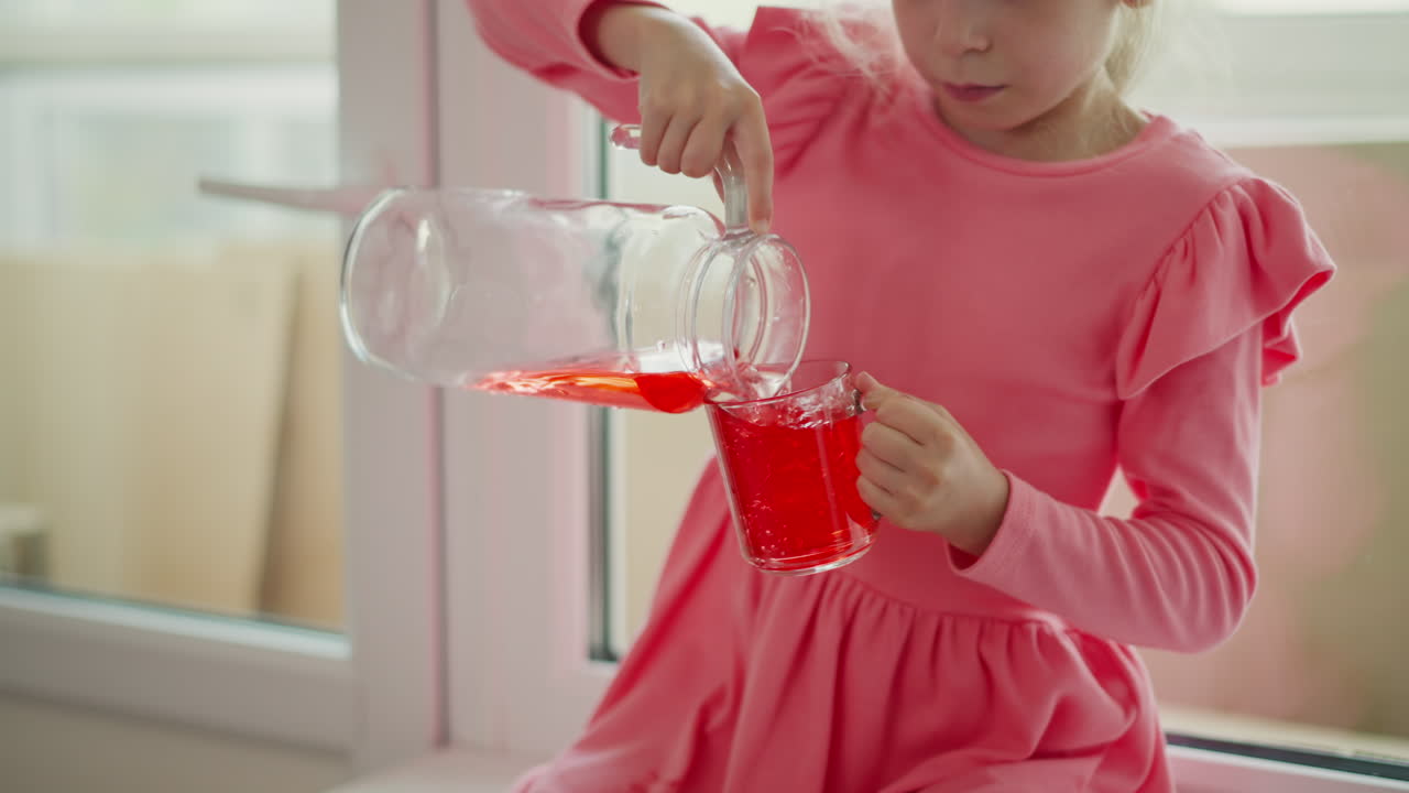 close up of young girl wearing pink dress sitting by window pouring red juice from glass jar into clear mug spilling drink on dress indoors playful cheerful focused casual refreshing childhood moment