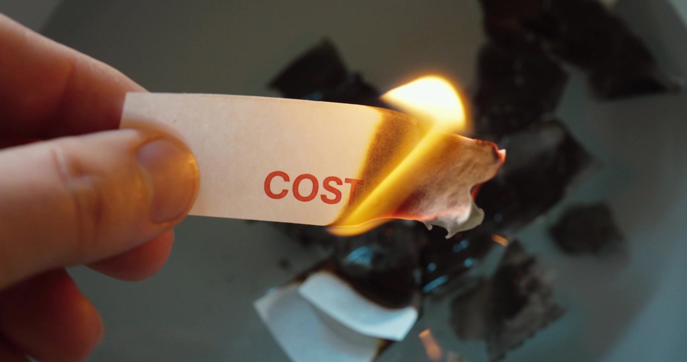 Ripresa ravvicinata al rallentatore di una striscia di carta con la parola “Costi” che brucia in una fiamma, a simboleggiare spese, pressione finanziaria, tagli al budget, inflazione, risparmi, rischio aziendale e sfide economiche.