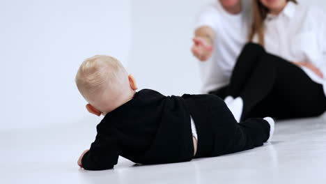lovely blond baby boy wearing black suit crawls by the floor. cheerful parents sit behind at backdrop. white background.