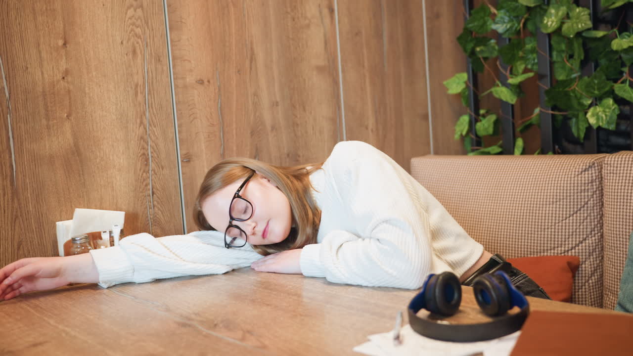 Young lady wearing white sweater removes headphone and leans her head forward in moment of fatigue at wooden table with drawing papers, pen, and book, indoor setting with leafy background