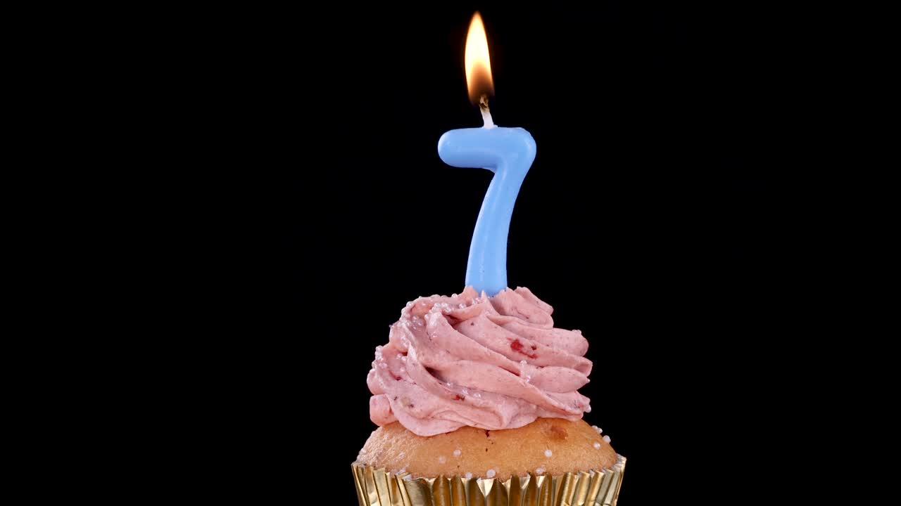 A single cupcake with pink frosting and a lit blue number seven candle stands against a black background, illuminated by the candle’s flame. Static camera, studio lighting