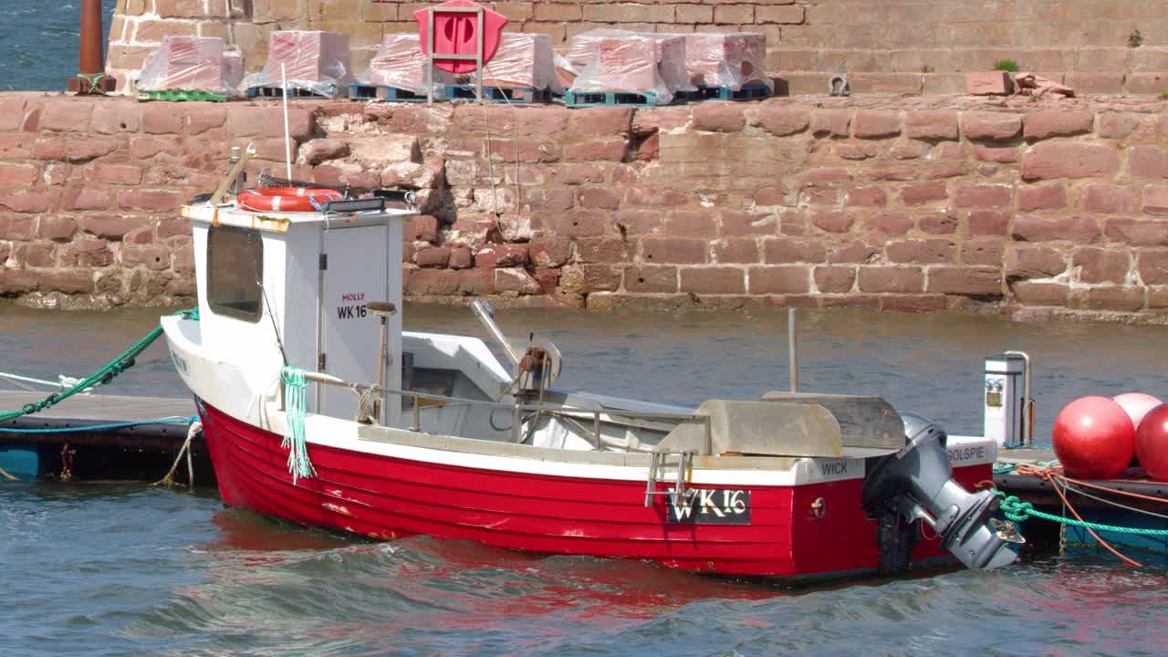 Red fishing boat moored at stone harbour wall, midday sun, gentle water movement, static camera