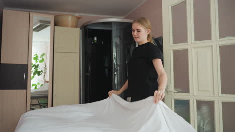 blonde woman spreads white sheet on bed in organized room featuring wall mirror and toilet sink, her motion captured mid-air with fabric floating gently, reflecting care and calm in domestic space