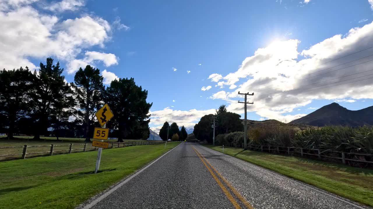 Vehicle travels scenic country road, passing fields, trees, and mountains under bright daylight sky
