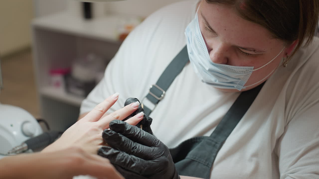 Close-up of focused nail technician in black gloves carefully working on customer fingernails with nail tool during manicure session, moving from one nail to the next in clean salon environment