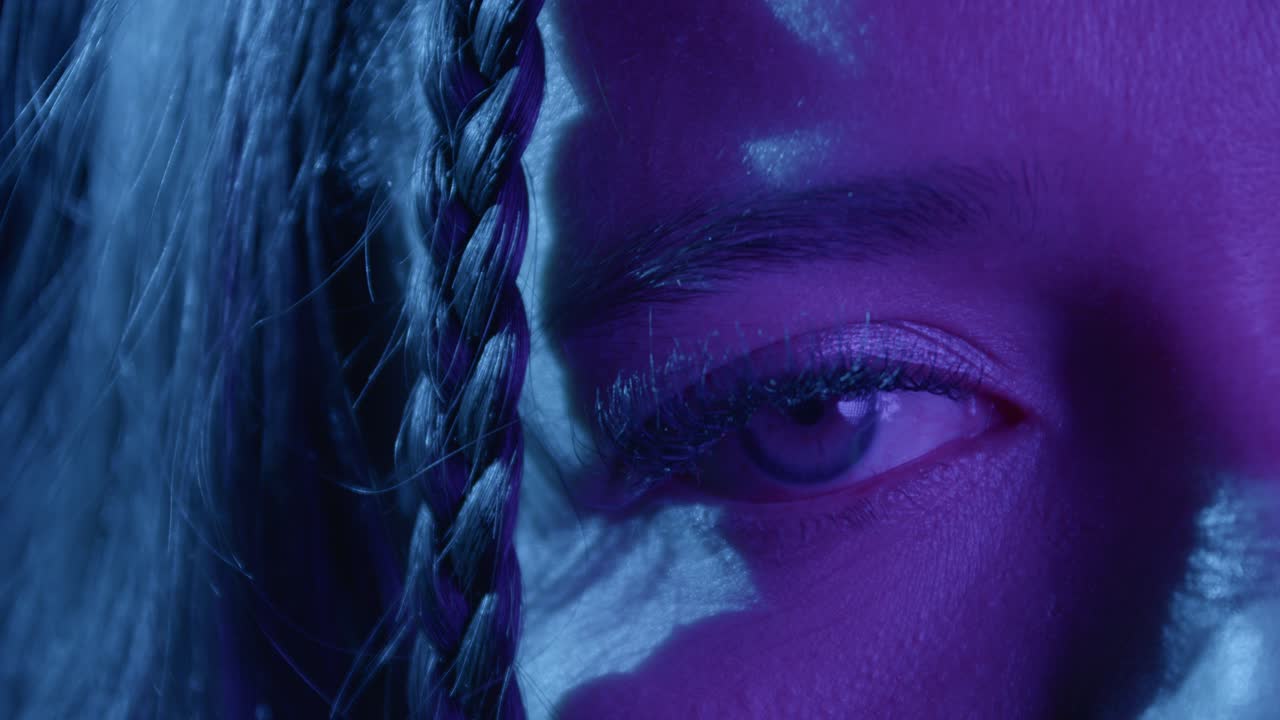 Detailed close-up shot of an eye framed by braided hair. The vivid blue and purple lighting creates a mysterious, dramatic mood, emphasizing texture and depth in the composition
