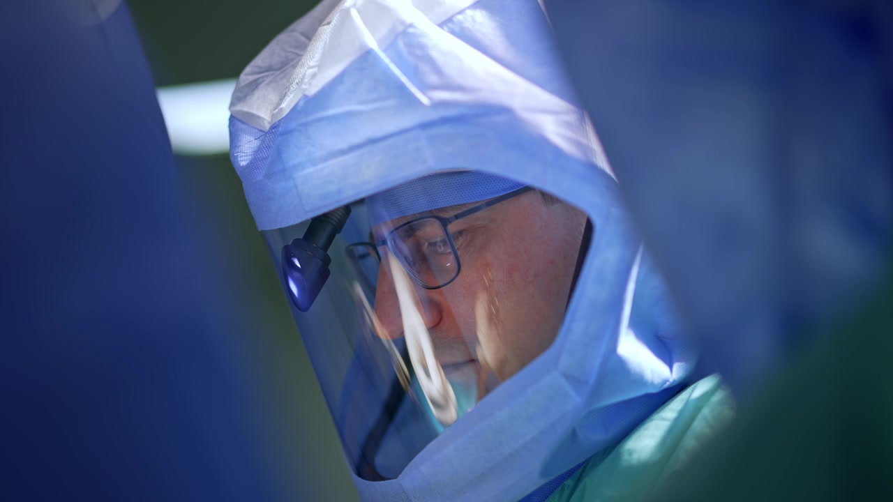 Chirurgo maschio caucasico di mezza età con occhiali, casco protettivo e lampada frontale. Ritratto ravvicinato di un medico impegnato in un intervento chirurgico.