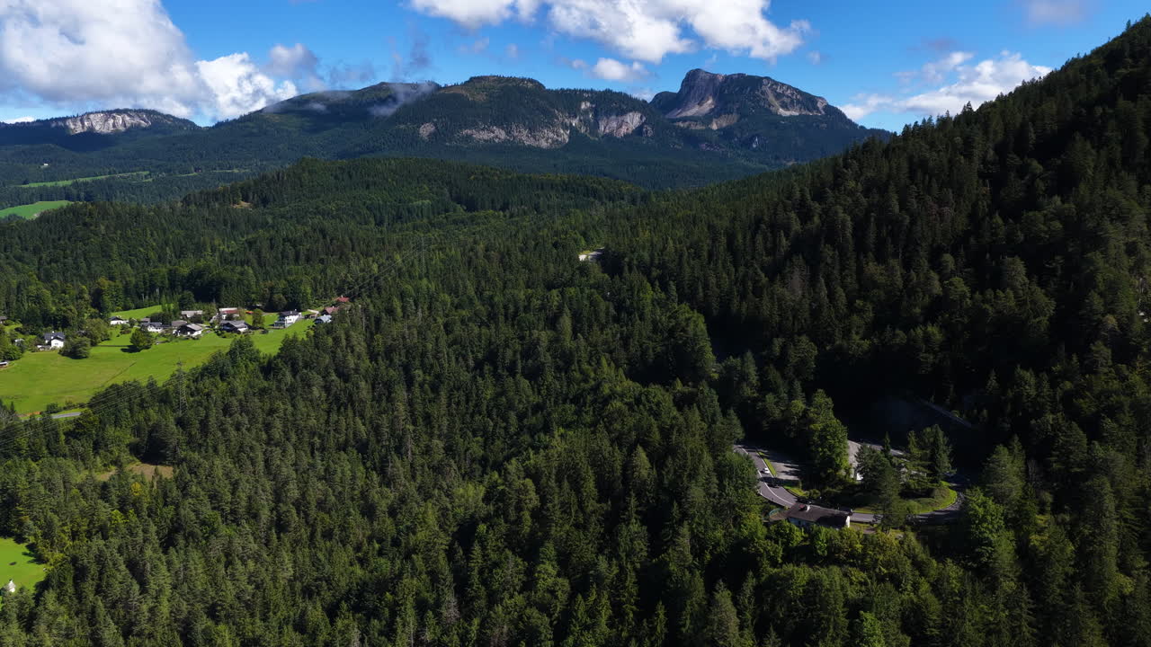 Cinematic aerial drone shot of dense alpine forest valley with rural houses and dramatic mountain ridge under blue sky. Scenic European alpine landscape