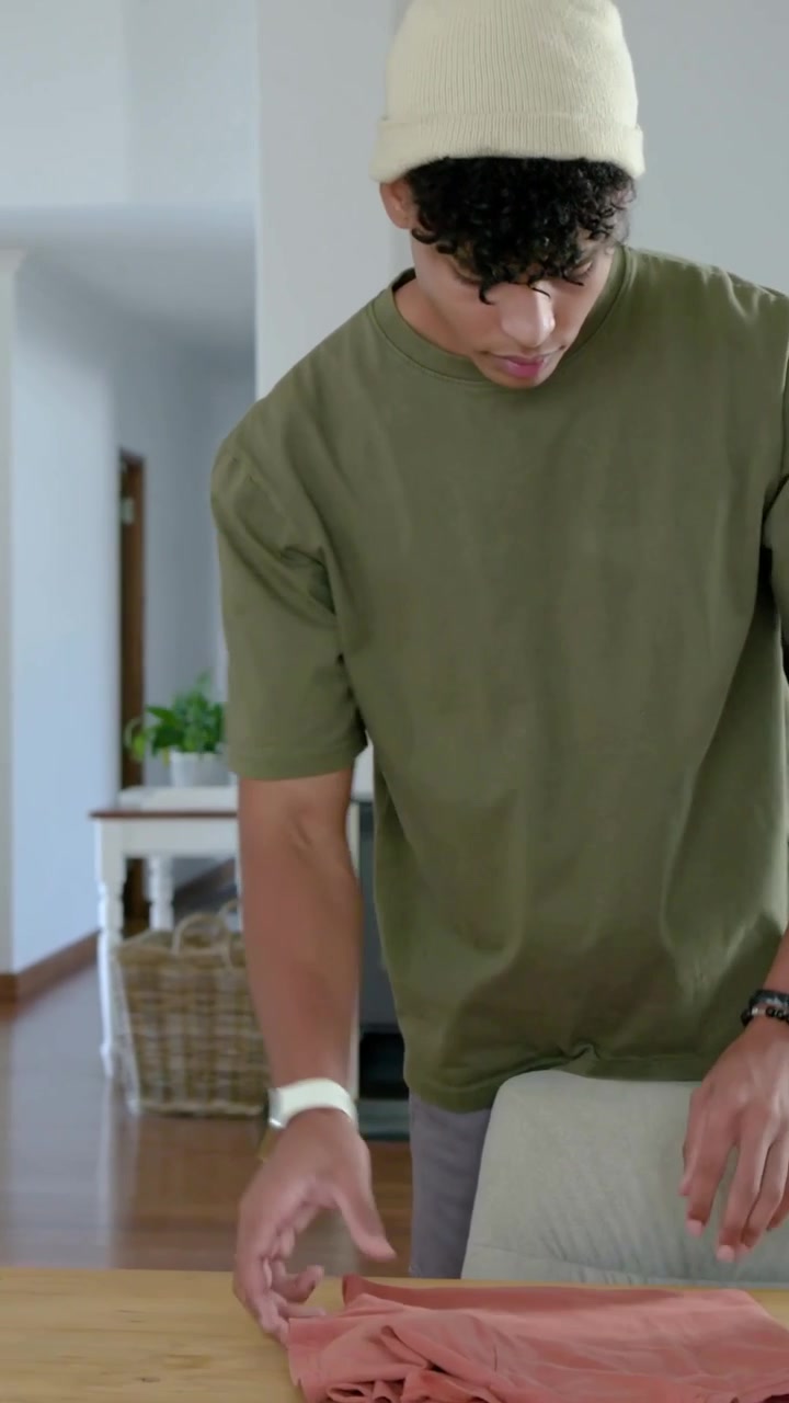 Vertical video: Picking rust shirt, man in olive tee checking, folding at home, camera panning left
