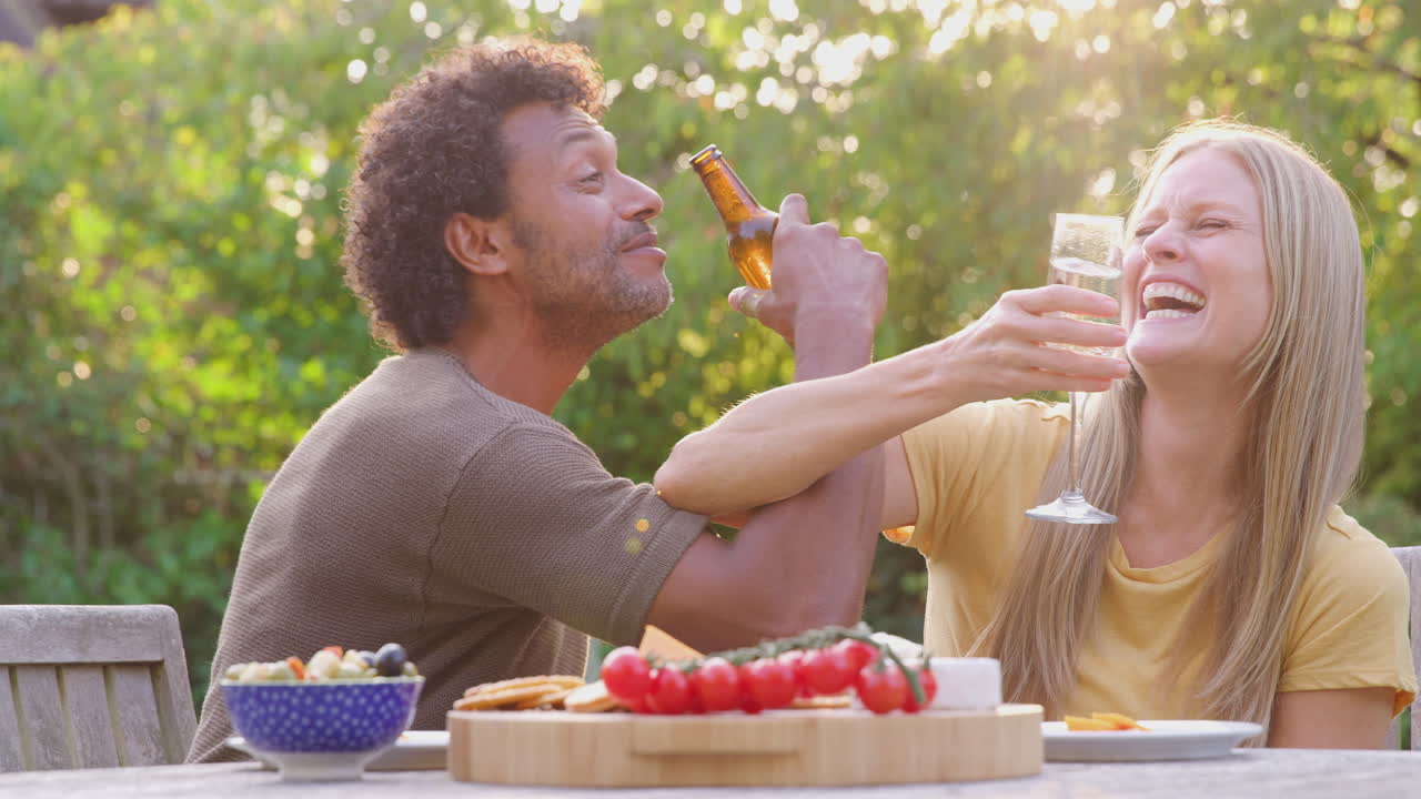 Volwassen stel arm in arm vierend met champagne en bier aan tafel in de tuin met hapjes
