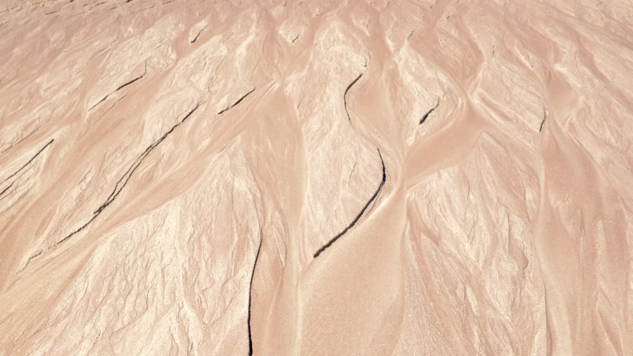 Water flows slowly down a beach and into teh sea creating beautiful, temporary patterns in the sand which branch as the water slowly erodes the sand, glistening in the sunlight.