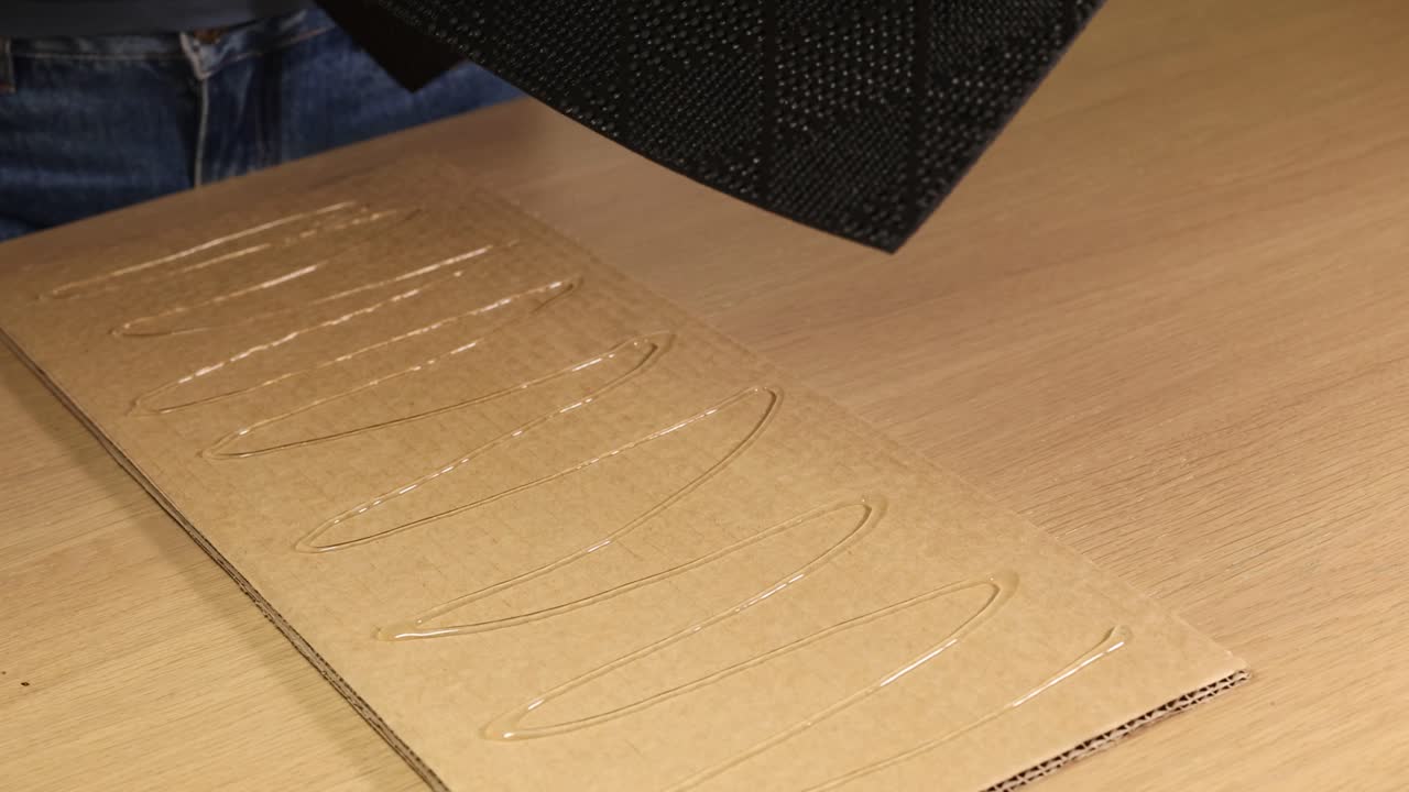 Hands pressing a textured rubber mat onto a glue-coated cardboard surface. Bright lighting, close-up shots, and steady camera focus