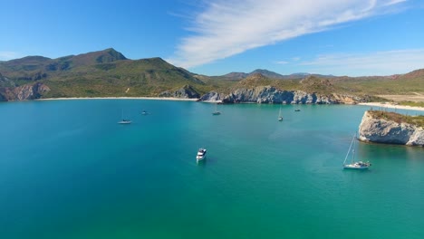 Schwenk-Luftaufnahme-Von-Segelyachten,-Die-In-Einer-Bucht-Vor-Anker-Liegen,-Mit-Einer-Felsigen-Insel,-Die-An-Einem-Wunderschönen,-Sonnigen-Sommertag-Mit-Blauem-Himmel-Durch-Die-Mitte-Taucht