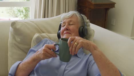 Kaukasische-Seniorin-Trägt-Kopfhörer-Und-Hört-Musik,-Während-Sie-Zu-Hause-Eine-Kaffeetasse-In-Der-Hand-Hält