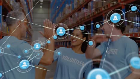 Animation-of-network-of-icons-over-three-diverse-volunteers-high-fiving-each-other-at-warehouse