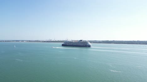 a vast cruise ship glides across the water, with industrial structures visible on the distant shore under a clear sky.