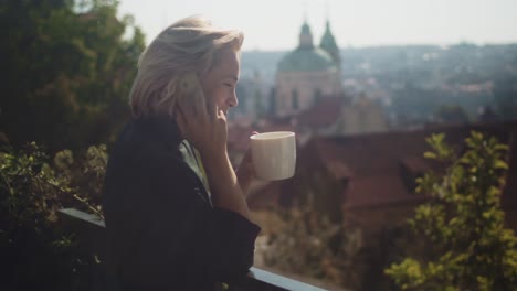 Mujer-De-Negocios-En-El-Balcón-Riéndose-Cuando-Habla-Por-Teléfono-Mientras-Bebe-Una-Taza-De-Café-Caliente