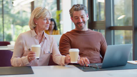 female businesswoman brings takeaway coffee for male colleague as they have meeting with laptop