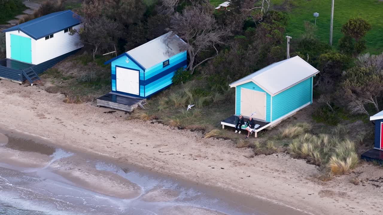 Drone pans above pastel beach huts along sandy shoreline, soft daylight, tranquil coastal atmosphere
