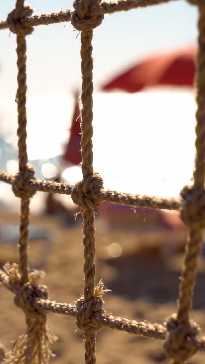 Un Primo Piano di una Recinzione di Corda Annodata che Incornicia una Vista sulla Spiaggia Illuminata dal Sole con Ombrelloni Vivaci sullo Sfondo, Creando un'Atmosfera Estiva Serena
