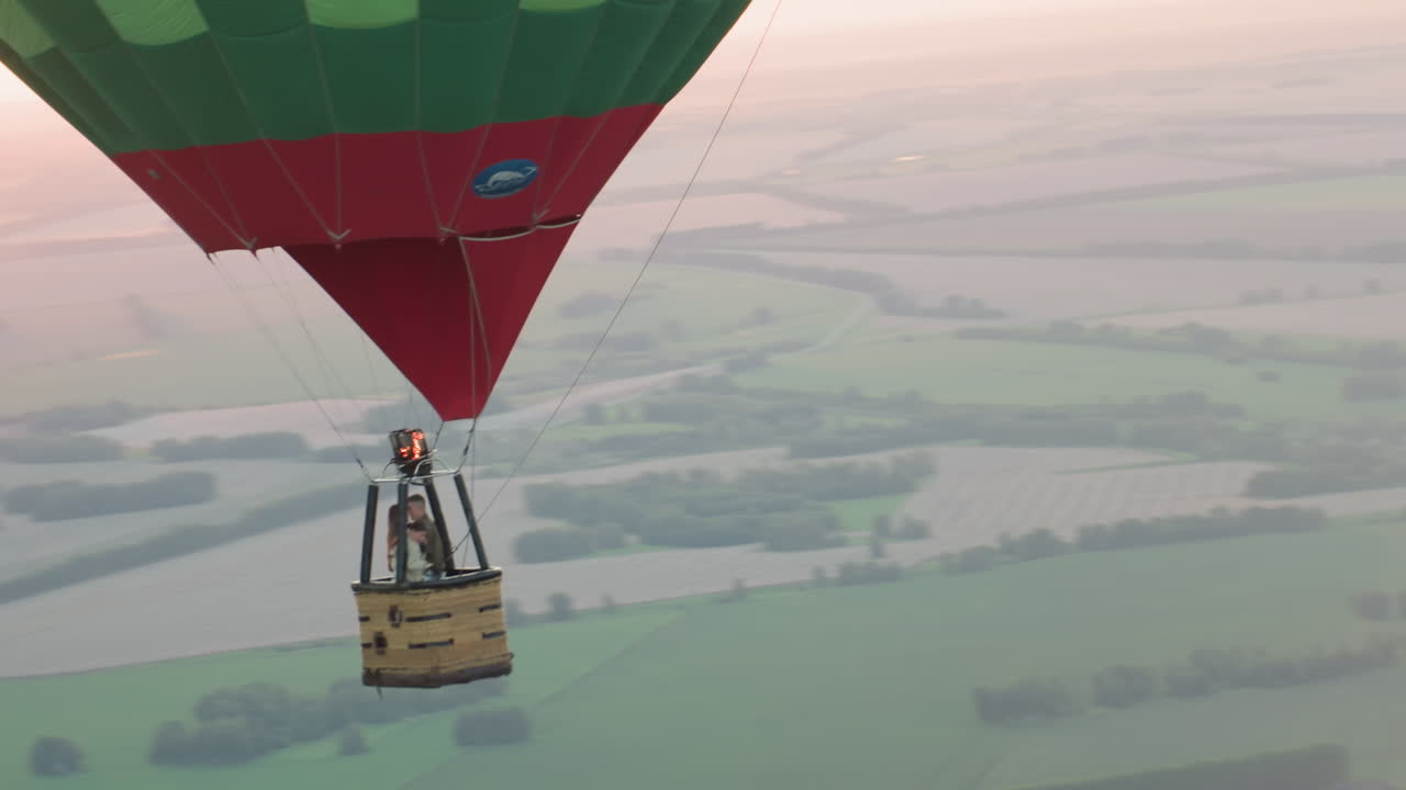 couple passengers riding hot air balloon above patchwork farmland at sunset with pilot igniting burner flame to ascend vessel drifting over green fields captured from aerial perspective