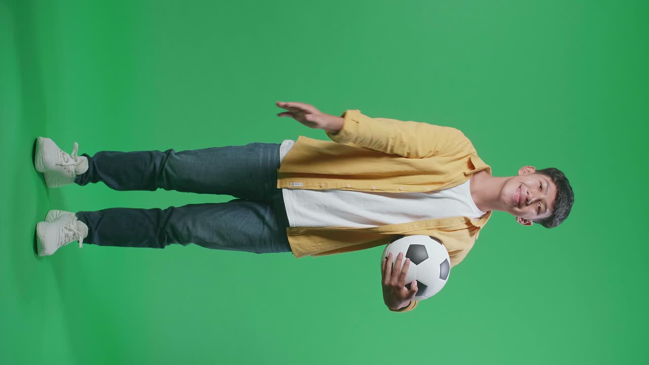 cuerpo lleno de niño asiático con una pelota soplando besos y sonriendo a la cámara mientras aplaude el fútbol en el fondo de la pantalla verde