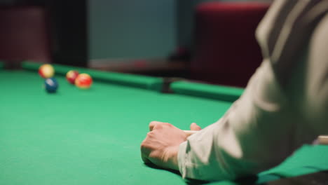 close-up view of player s hand in white shirt taking precise shot on green pool table. cue stick positioned, white ball rolling forward. blurred background with soft lighting