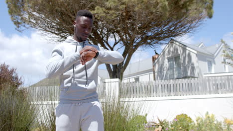 happy african american man jogging and using headphones and smartwatch on sunny day, slow motion