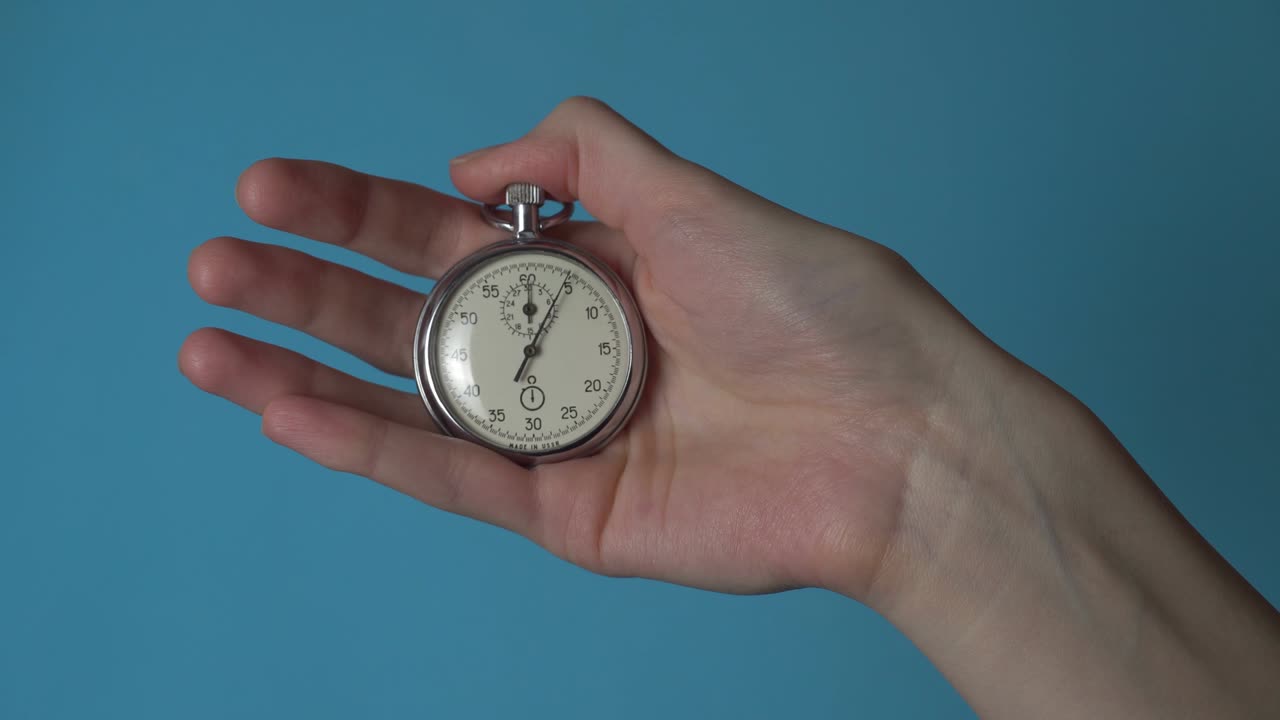A woman's hand holds an analog stopwatch on a blue chromakey screen.