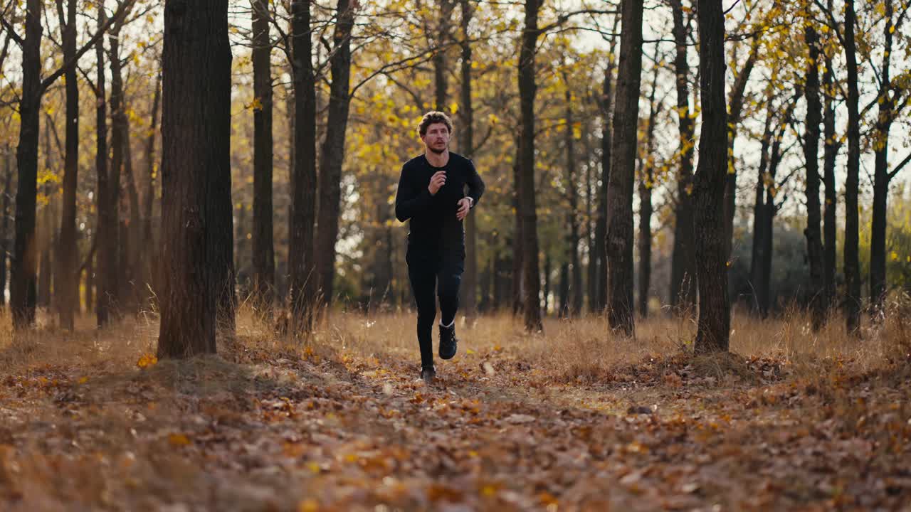 A man in a black sports uniform with curly hair and a beard looks at his watch and then runs through the fallen leaves in the autumn forest during his morning jog