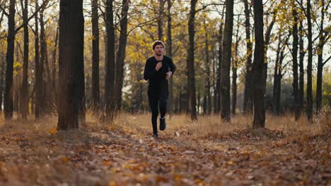 A-man-in-a-black-sports-uniform-with-curly-hair-and-a-beard-looks-at-his-watch-and-then-runs-through-the-fallen-leaves-in-the-autumn-forest-during-his-morning-jog