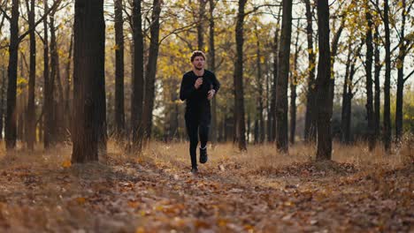 a man in a black sports uniform with curly hair and a beard looks at his watch and then runs through the fallen leaves in the autumn forest during his morning jog