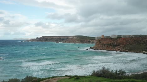 AERIAL:-Ghajn-Tuffieha-Bay-on-a-Gloomy-but-Bright-Day-with-Sea-Roaring-and-Splashing-into-Shore