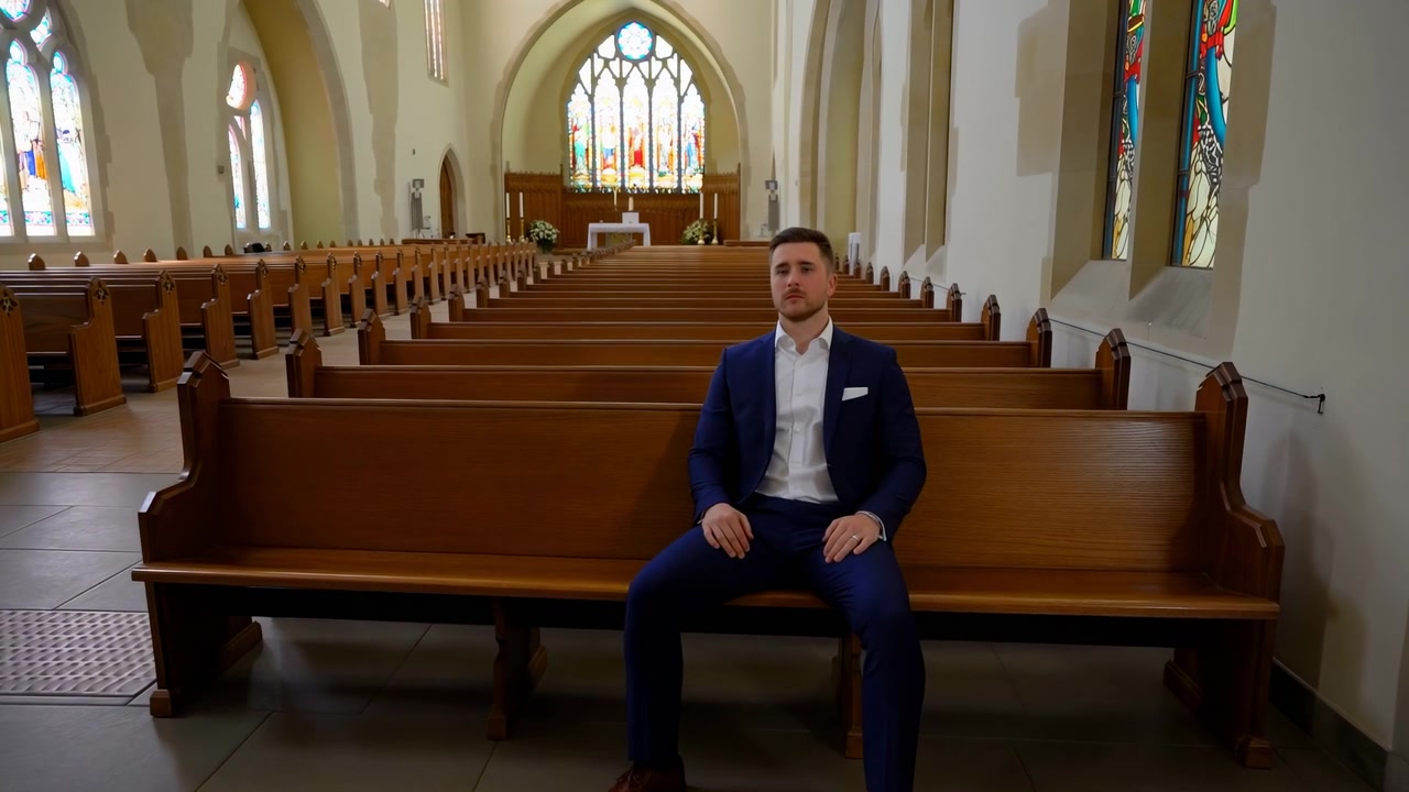 Young man in dark blue suit and white shirt sits on front wooden pew inside traditional church nave, hands resting on thighs while turning head and torso toward camera under colorful stained glass