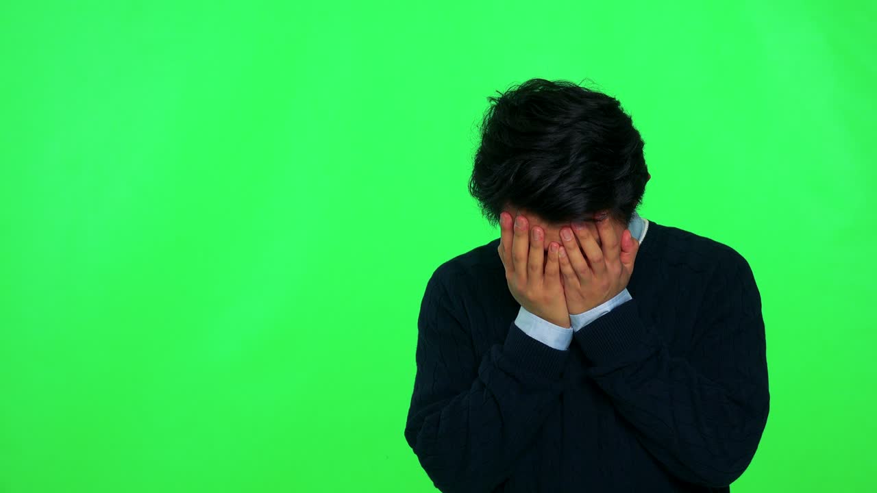 A young Asian man covers his face with his hands, then stops and looks around, confused - green screen studio