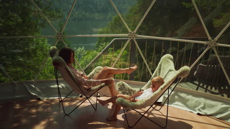 lady in home clothes sits with son on glass balcony looking at wide window. mother and son relax enjoying view of calm lake and forest. illustration of fantastic nature