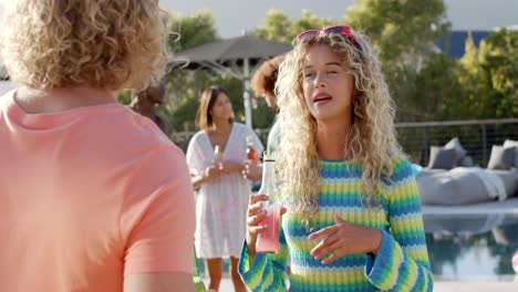 Happy-caucasian-couple-with-drinks-talking-at-sunny-pool-party-with-diverse-friends,-slow-motion