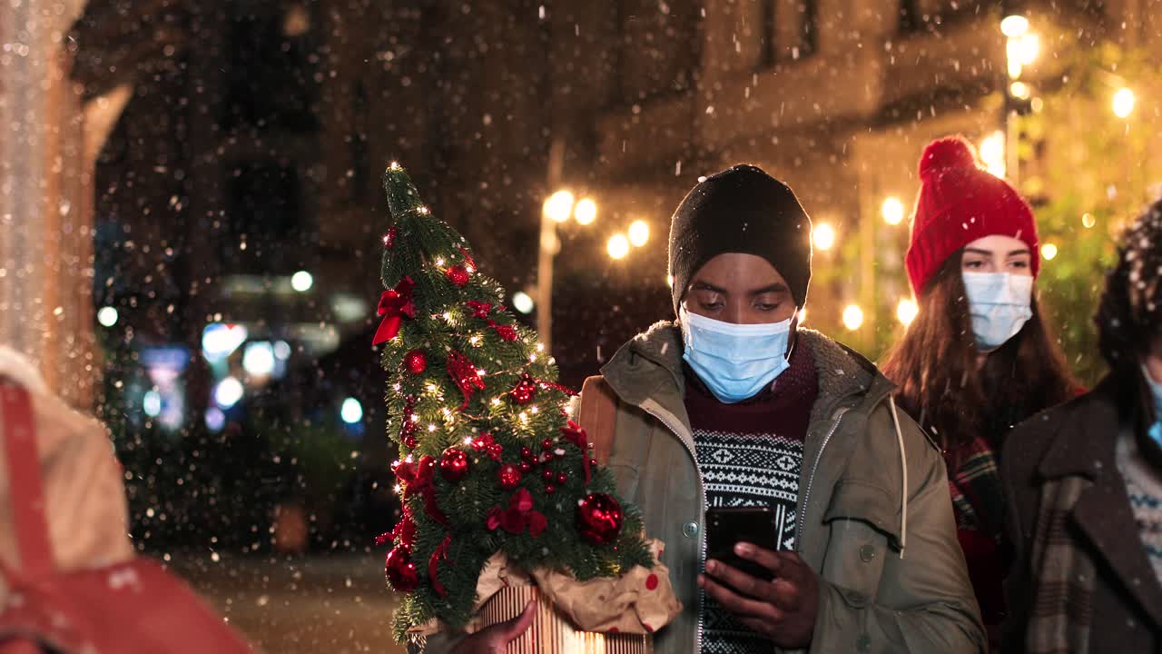 크리스마스에 눈이 내리는 동안 스마트폰으로 입력하고 거리에서 크리스마스 트리를 들고 있는 아프리카계 미국인 남자의 초상화