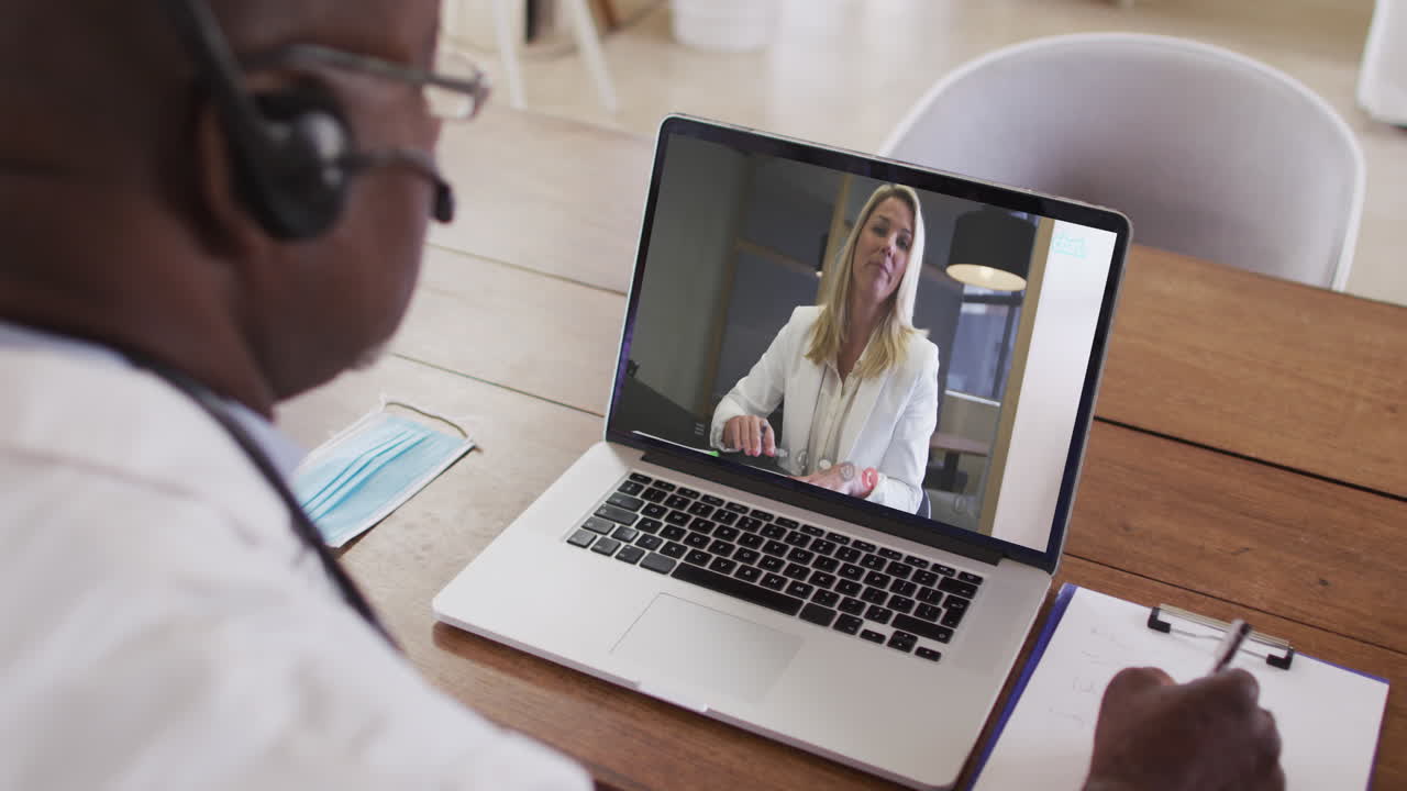 Afroamerikansk forretningsmand sidder ved skrivebord, bruger bærbar computer og har videoopkald med en kvindelig kollega.