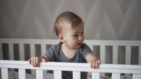 lovely kid standing up in his cot holding by the handrails. beautiful child looking around after waking up. blurred backdrop.