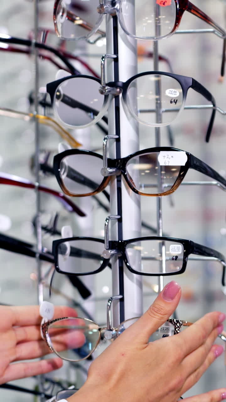 Young female choosing eyeglasses in the store. Hands of a woman customer spinning the stand with fashionable spectacles at optics. Vertical video