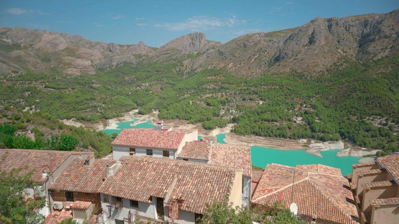 Case con tetti in terracotta si affacciano su acqua turchese che serpeggia attraverso una valle montuosa boscosa, con scogliere aspre e pendii verdi che riempiono lo sfondo sotto una luce diurna chiara e brillante, creando un'atmosfera di viaggio tranquilla