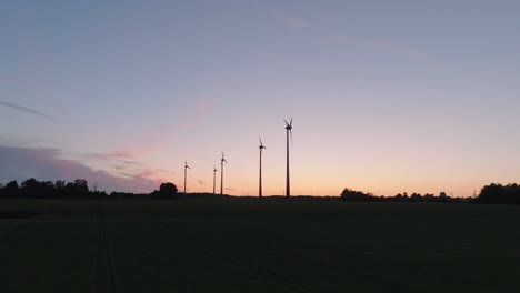 Aerial-establishing-view-wind-turbines-generating-renewable-energy-in-a-wind-farm,-evening-after-the-sunset-golden-hour,-countryside-landscape,-high-contrast-silhouettes,-drone-shot-moving-forward