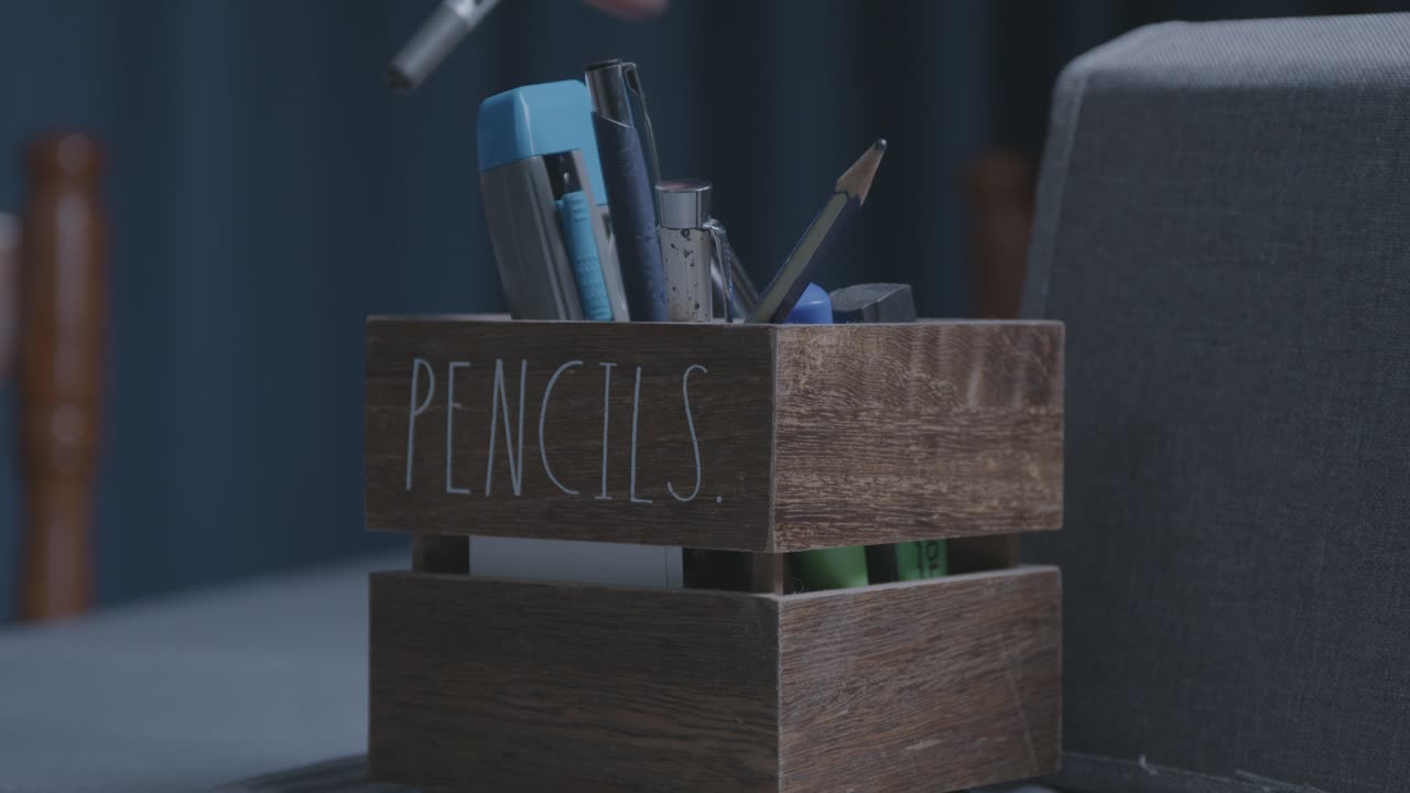 A cinematic close-up of a rustic wooden pencil holder on a desk. A hand reaches in to select and take out a pen. Ungraded LOG for a professional, customizable look